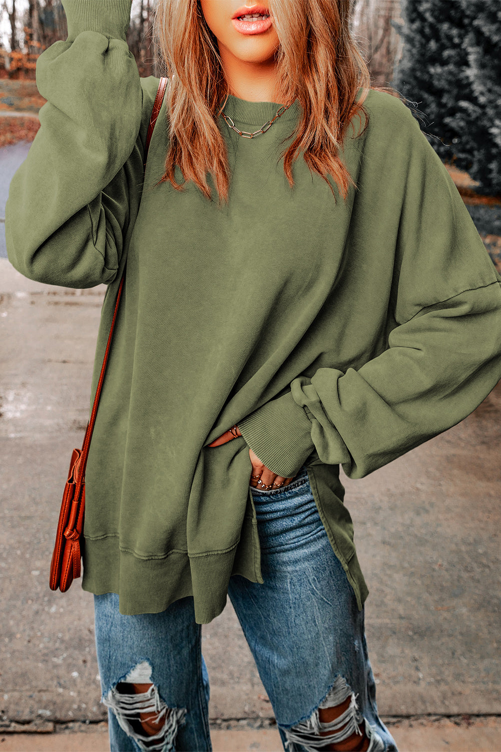 A person with wavy, light brown hair, wearing a Green Drop Shoulder Ribbed Trim Oversized Sweatshirt made of skin-friendly fabrics, blue jeans, and holding a brown shoulder bag is walking outdoors on a concrete path.