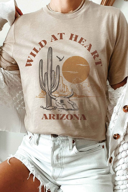 Woman wearing the WILD AT HEART ARIZONA Graphic Tee in white, which features the text "Wild at Heart Arizona" along with an illustration of a cactus, a sun, and a lounging skeleton. This tee is made from 100% cotton and offers unisex sizing for ultimate comfort and style.