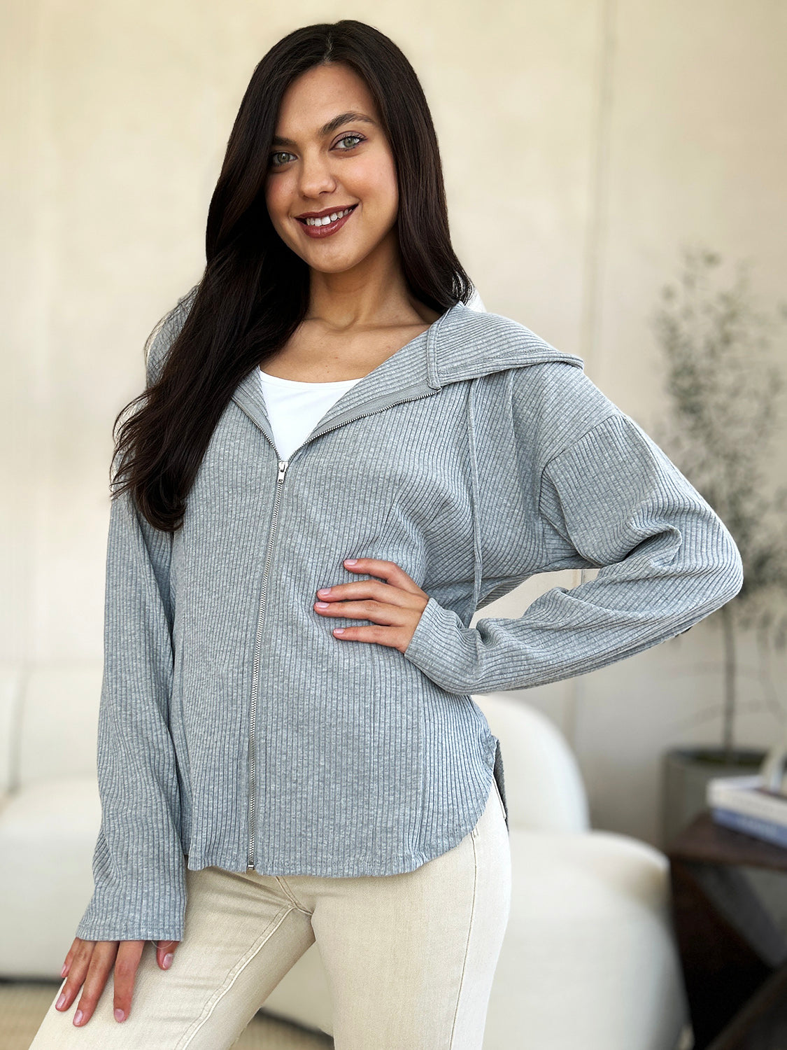 A woman in a trendy Culture Code Full Size Ribbed Zip Up Drawstring Hooded Jacket and light-colored pants stands indoors, smiling with her eyes closed and one hand resting on her chin.