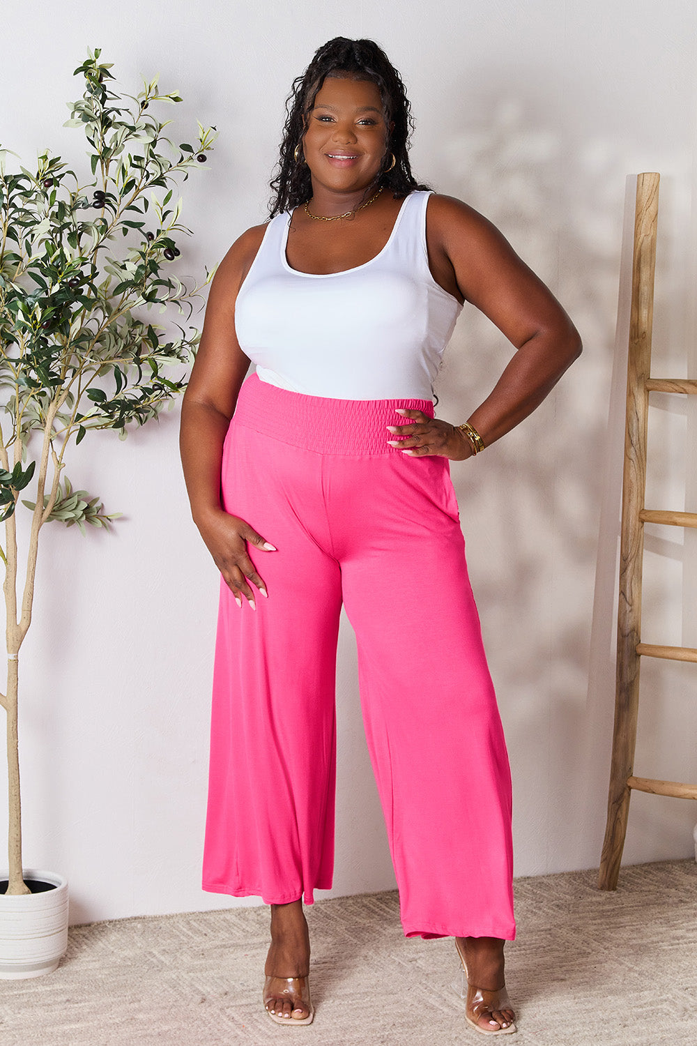 A curve model is wearing the Double Take Full Size Smocked Wide Waistband Wide Leg Pants in gray with hands in pockets, paired with white sneakers.