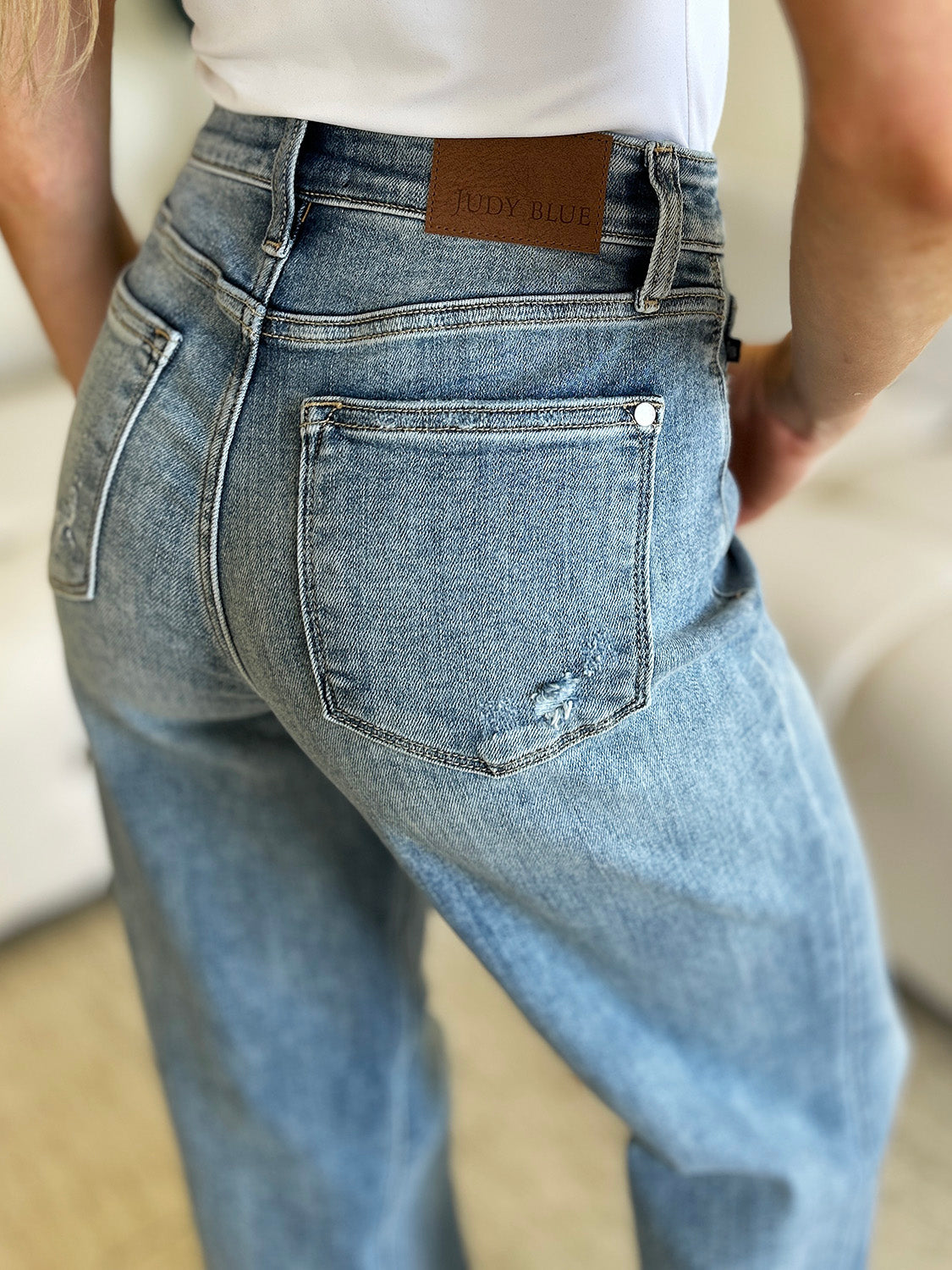 A person wearing Judy Blue Full Size High Waist Straight Jeans and white shoes is standing indoors. There is a white couch and a large plant in the background.