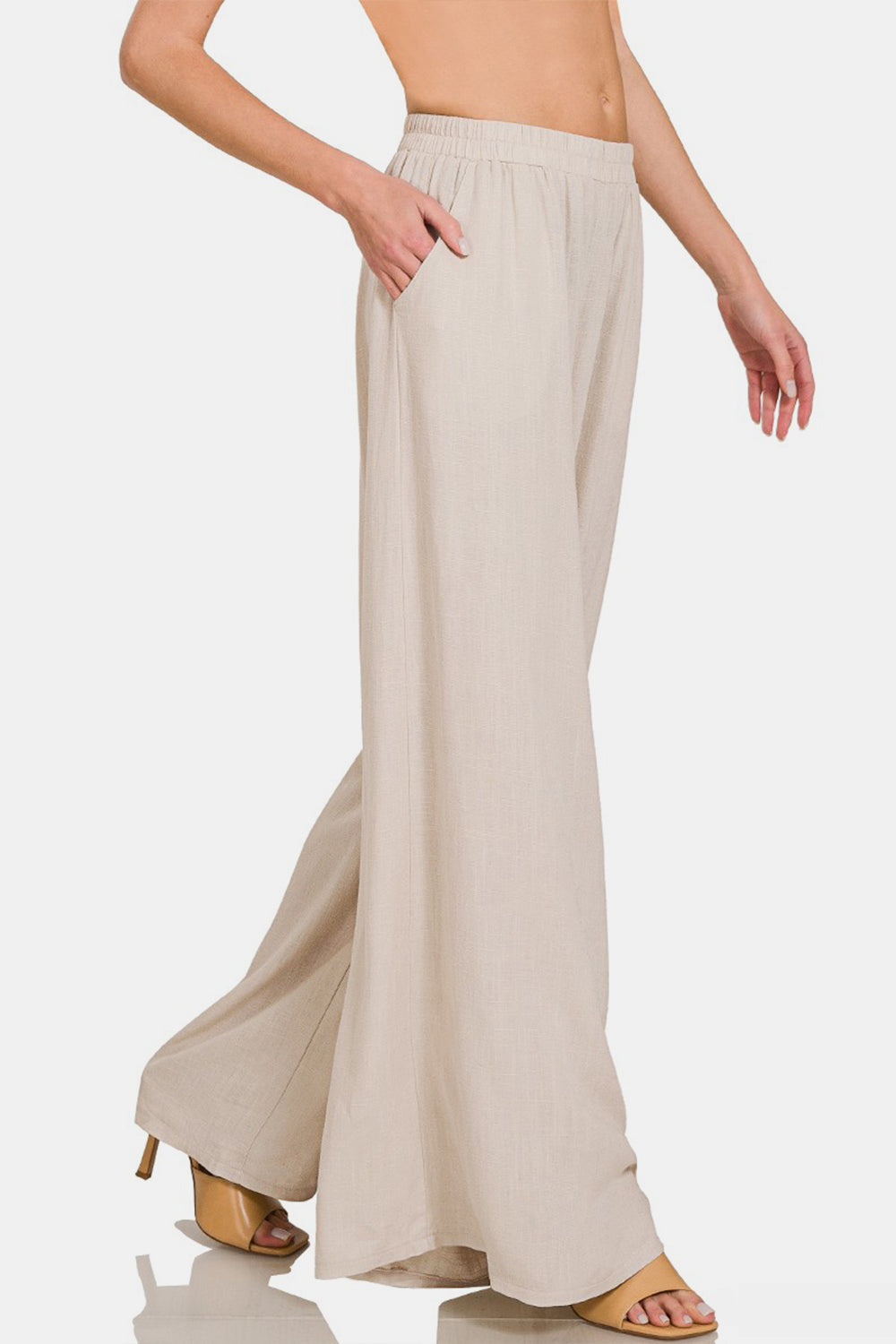 Person wearing Zenana Pleated Linen Blend Wide Leg Pants in beige with hands in pockets, shown from waist to feet. The pleated pants have an elastic waistband and flowy fabric. Model is also wearing light-colored open-toed sandals.