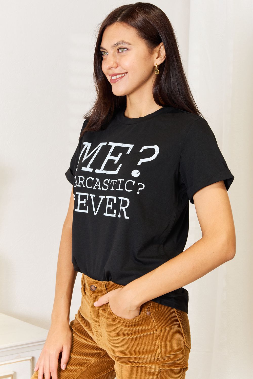 A woman with long dark hair smiles while wearing a trendy black Simply Love Letter Graphic Round Neck T-Shirt featuring the text "ME? Sarcastic? NEVER" and brown corduroy pants. She stands in a room with white walls and decor.