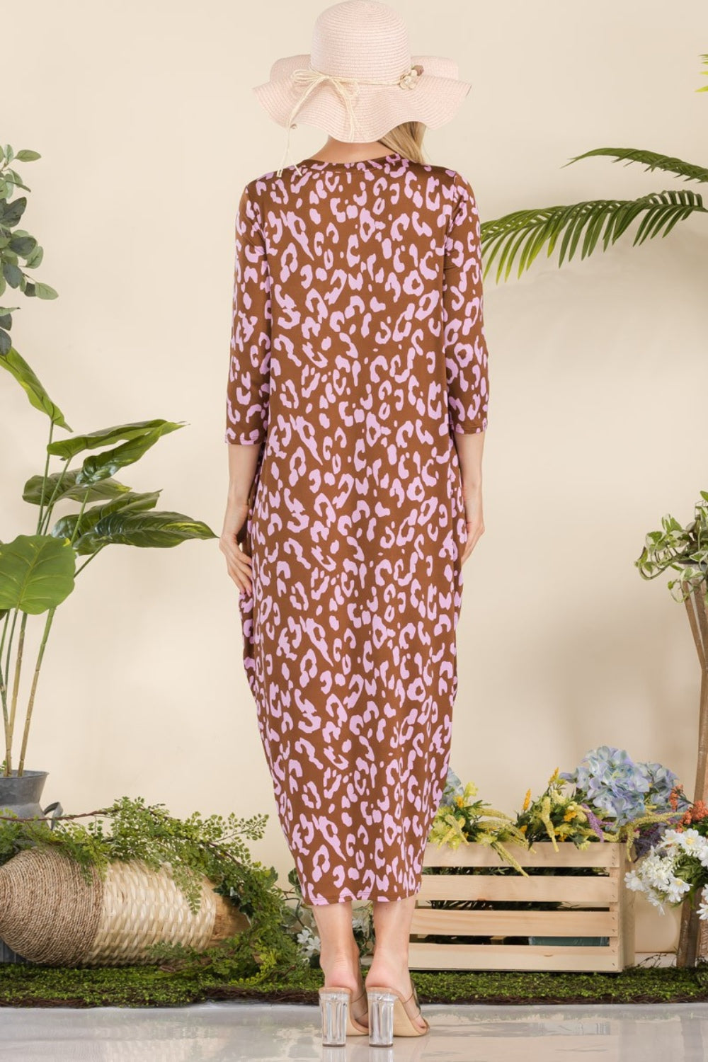 A woman in the Celeste Full Size Leopard Contrast Dress with pockets, featuring a brown and pink leopard print design and 3/4 sleeves, stands smiling among potted plants and wooden crates while wearing a wide-brim hat and sandals.