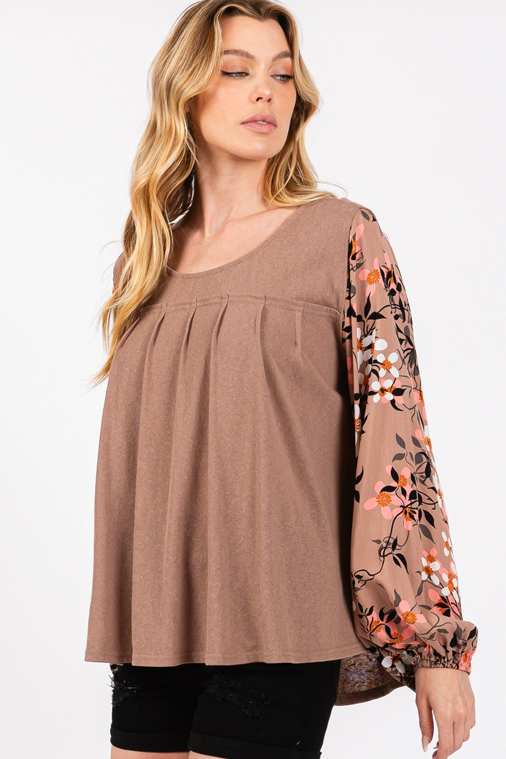 A person is wearing the SAGE + FIG Floral Long Sleeve Front Pleated Detail Blouse, paired with black shorts and a black hat, standing against a plain background.