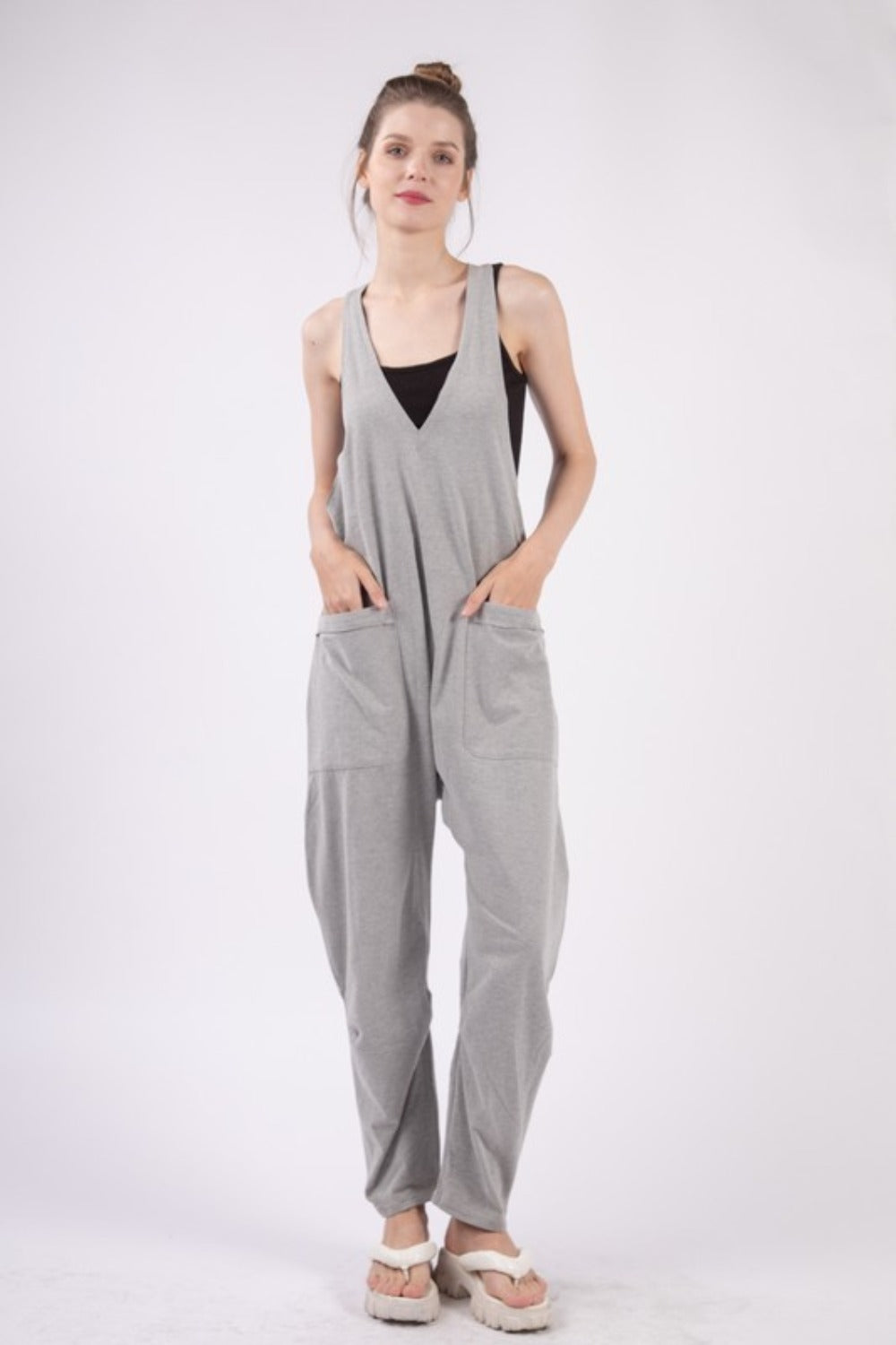 A person wearing the VERY J Plunge Sleeveless Jumpsuit with Pockets in gray and white sandals stands against a plain white background.