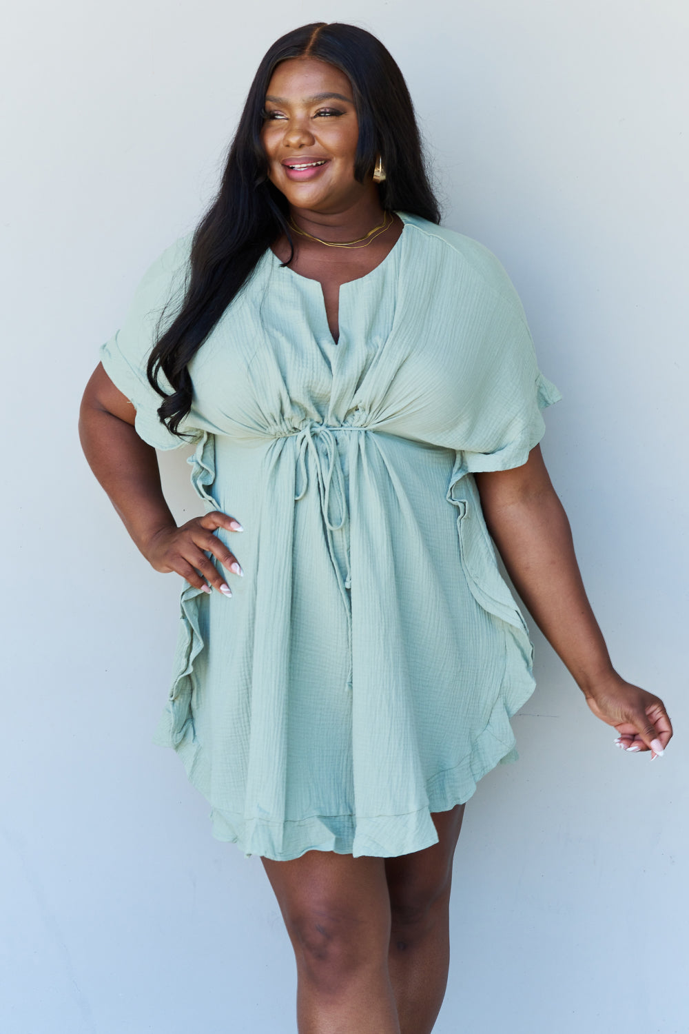 A woman is wearing the Ninexis Out Of Time Full Size Ruffle Hem Dress with Drawstring Waistband in Light Sage. Her long, dark hair falls gracefully as she smiles and looks to her left, standing against a plain background.