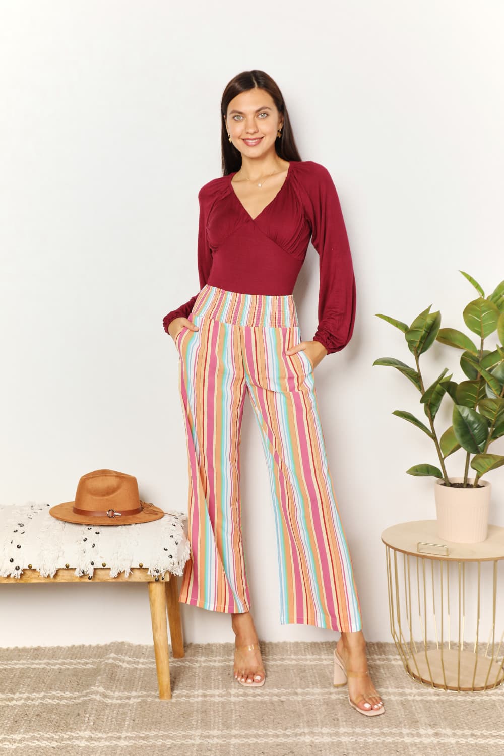 A person wearing Double Take Striped Smocked Waist Pants with Pockets and a burgundy long-sleeve shirt stands beside a wooden stool with a hat on it.