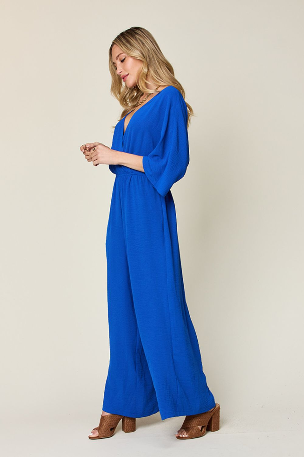 Person wearing the Double Take Full Size Surplice Wide Leg Jumpsuit with Pockets in royal blue, featuring wide legs, a deep V-neckline, and ruched detailing, posing with hands on hips against a plain light-colored background.