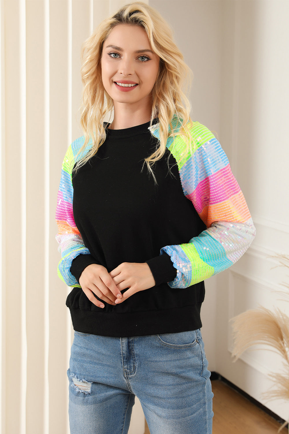 A person with shoulder-length hair is wearing a Black Sequin Color Block Raglan Sleeve Pullover Sweatshirt and blue jeans. They are facing away from the camera in a room with a potted plant.