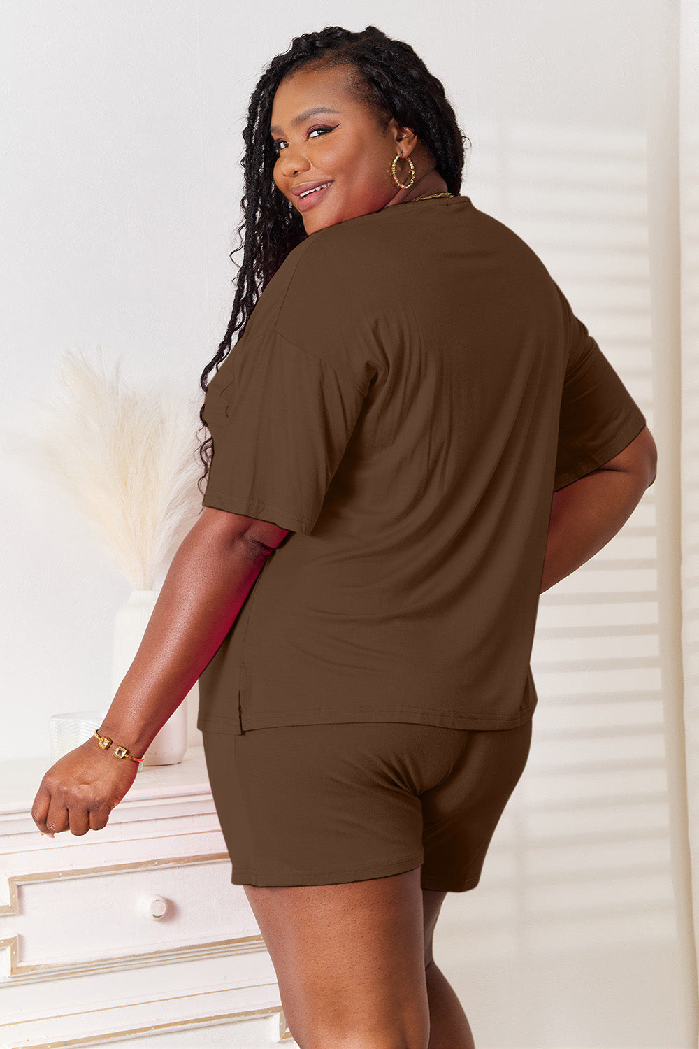 A woman is standing in a brightly lit room, wearing the Basic Bae Full Size Soft Rayon Half Sleeve Top and Shorts Set in a relaxed, stretchy turquoise fabric. She has one hand placed in her pocket and is smiling while looking slightly to the side.