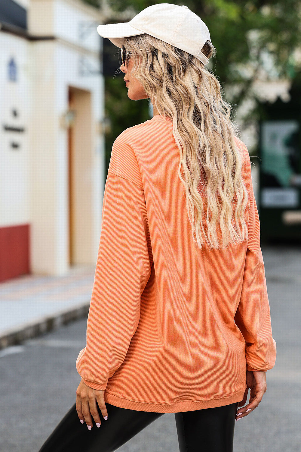 A woman with wavy blonde hair, wearing an Orange Corded SPICY GIRL Graphic Sweatshirt, black leggings, a beige baseball cap, and sunglasses, stands on a street facing away from the camera.