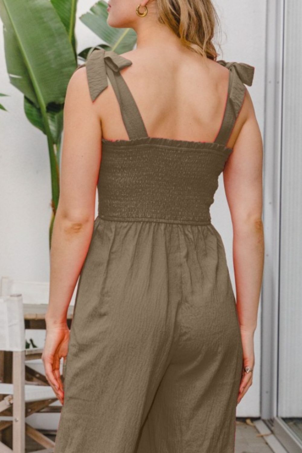 Clad in the elegant ODDI Full Size Bodice Smocked Sleeveless Jumpsuit, a woman strides confidently towards the camera on a patio beautifully decorated with plants and stylish furniture.