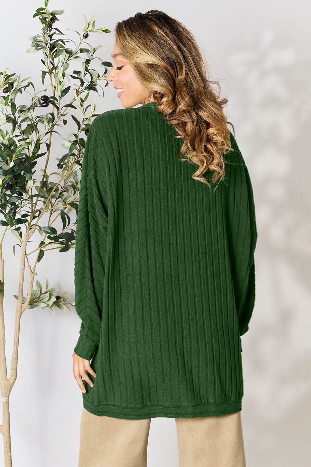 A person with wavy hair, wearing the Basic Bae Full Size Ribbed Cocoon Cardigan over a white top and beige pants, stands in front of a light-colored background with a leafy plant, showcasing an effortlessly basic style.