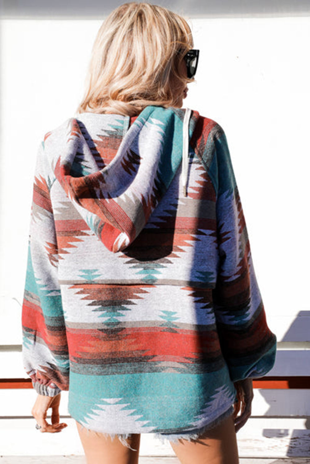 A woman with long blonde hair is standing outside, facing away from the camera. She wears a Multicolor Aztec Print Zipped Split Neck Hoodie and jeans. A rug and potted plant are visible in the background, adding to the cozy vibe of her super comfortable outfit.