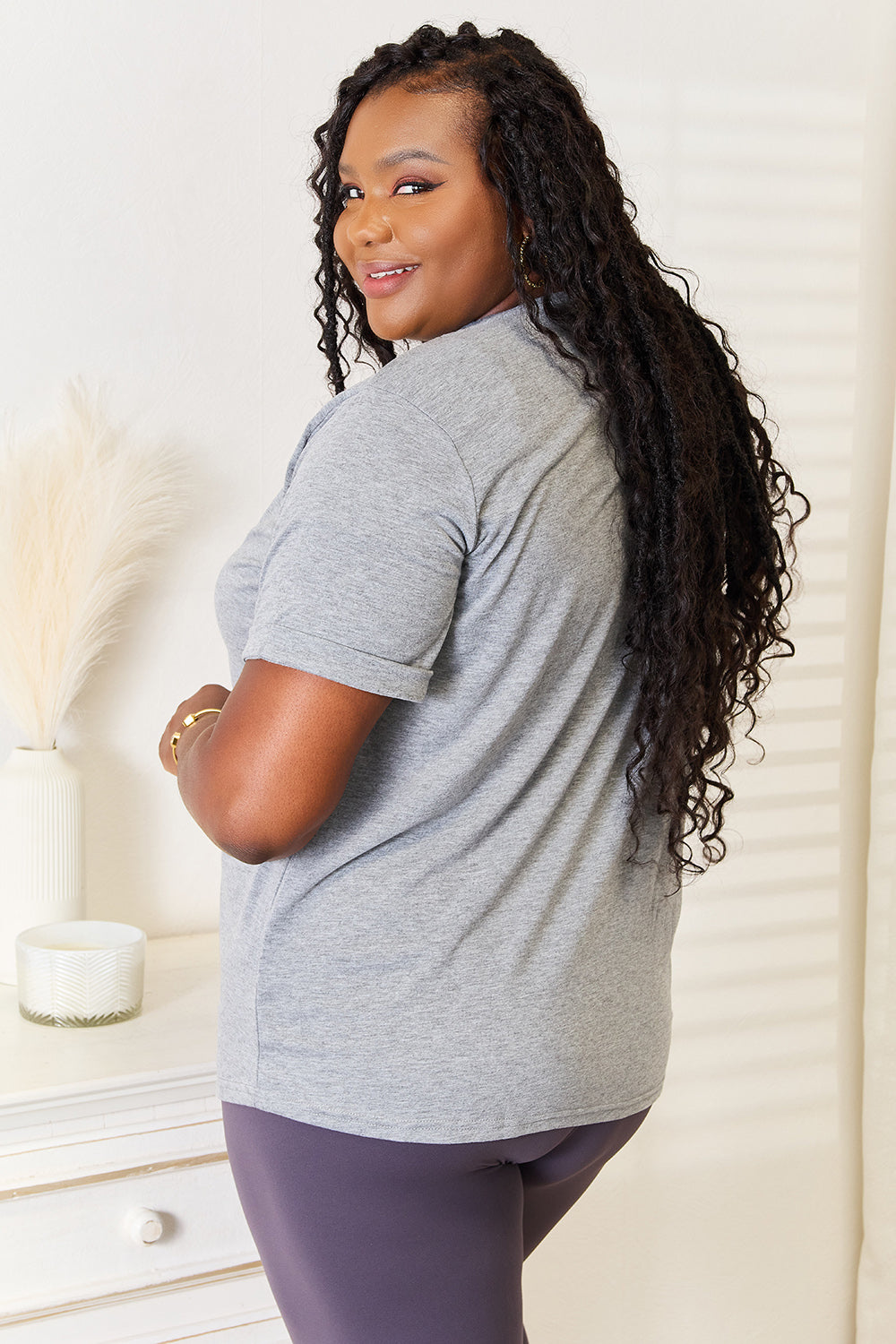 A woman with long hair stands indoors, smiling and touching her hair, wearing the trendy Simply Love Heart Graphic Cuffed Short Sleeve T-Shirt in gray.