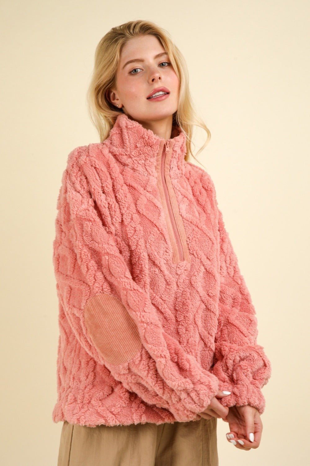 A person with long blonde hair wears a VERY J Fuzzy Fleece Half Zip Cable Pattern Sweatshirt in pink, paired with beige pants, standing against a plain light background. The sweatshirt is adorned with textured details and elbow corduroy patches.