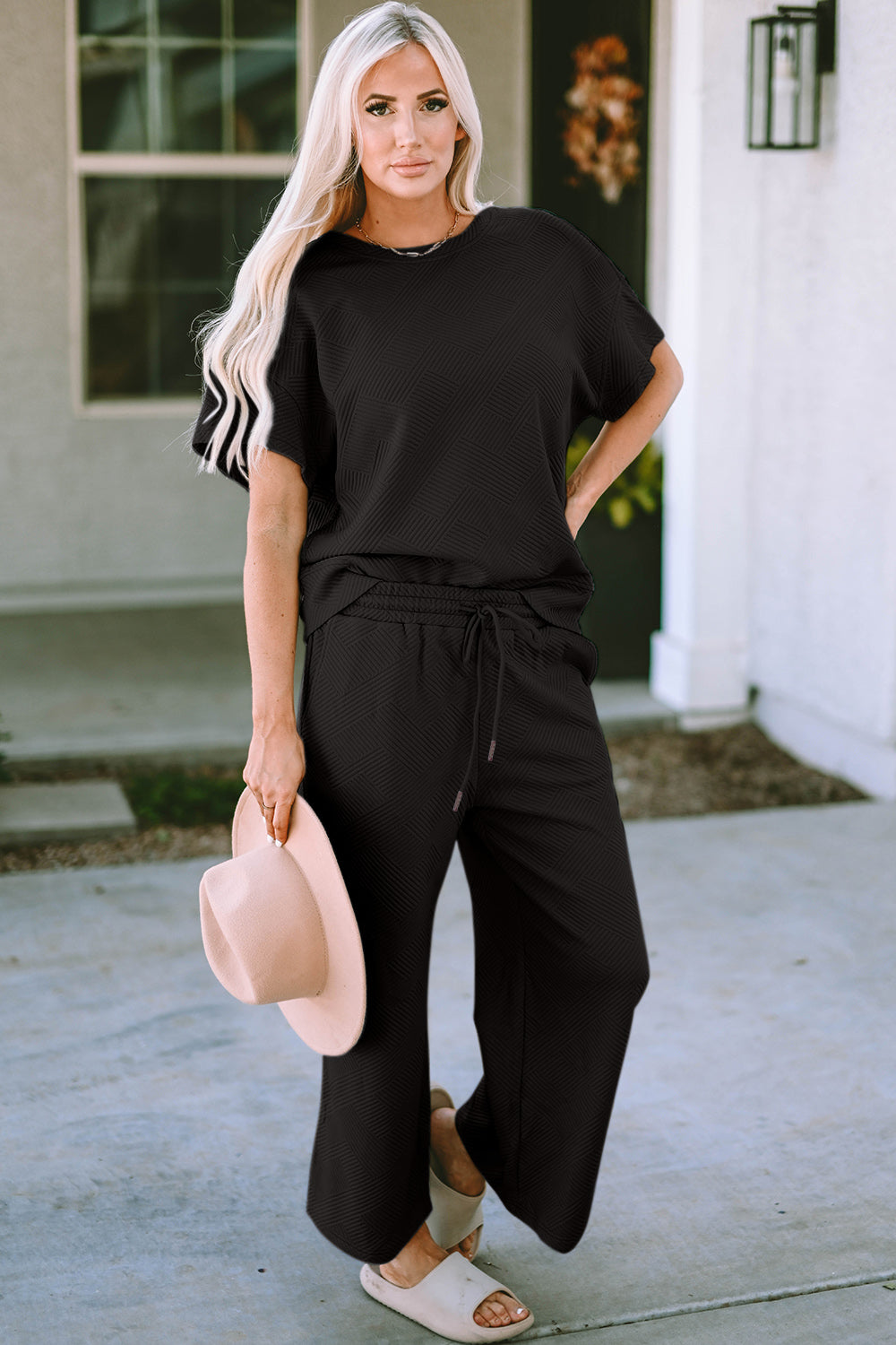 A woman with long hair smiles while standing outside a building, dressed in the Double Take Full Size Texture Short Sleeve Top and Pants Set, featuring a matching beige two-piece ensemble. She wears slides and holds a hat in her right hand. Her outfit, slightly stretchy for added comfort, is perfect for machine wash cold.