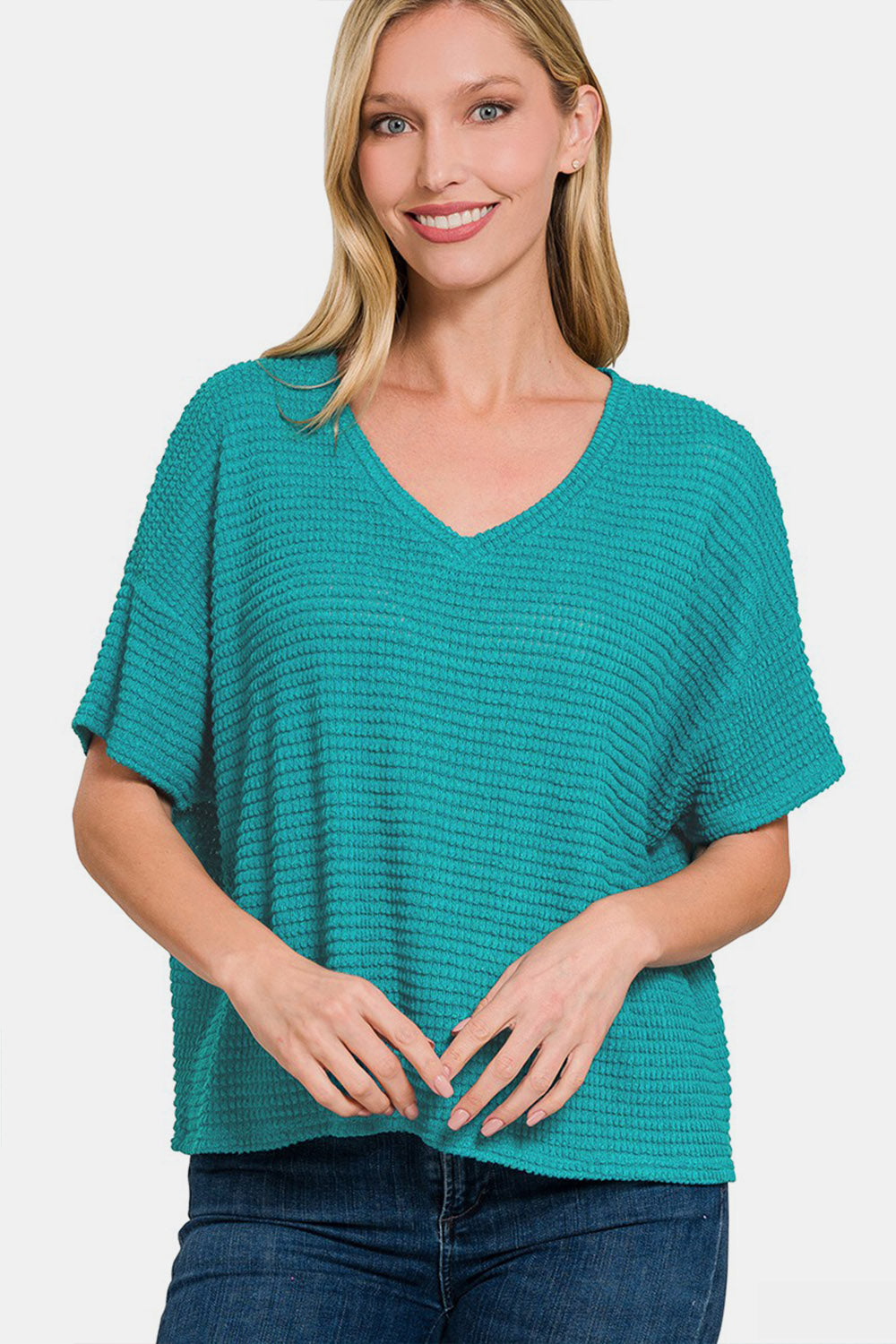 A person wearing a Zenana Drop Shoulder Short Sleeve Jacquard Knit Top in teal, paired with blue jeans, smiling against a plain white background.