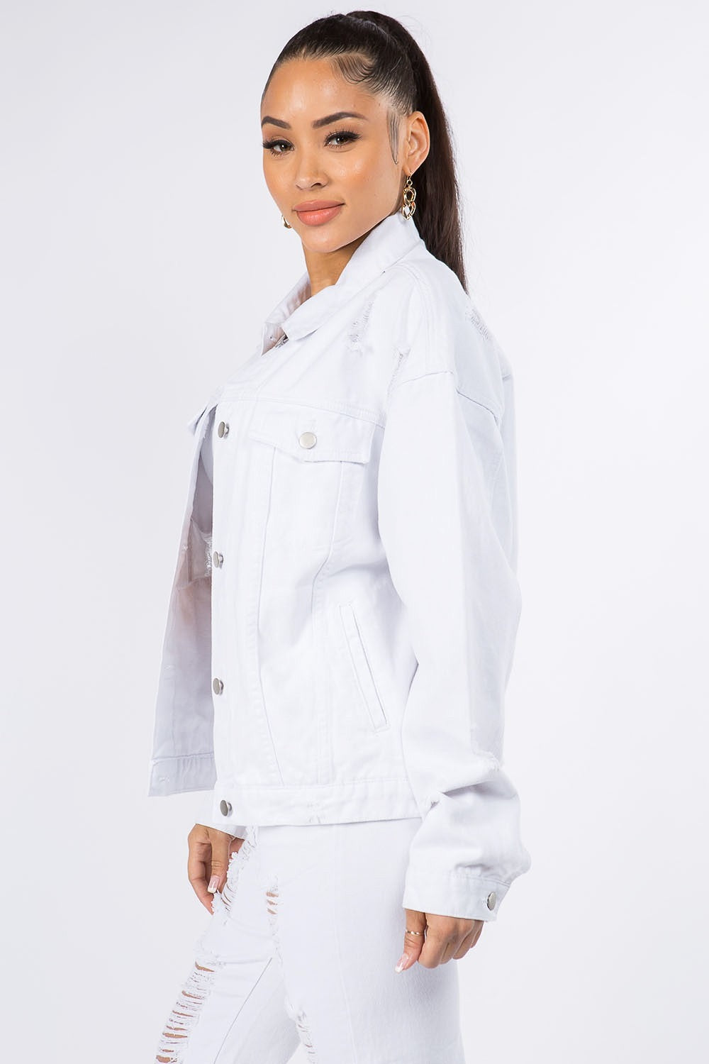 A woman wearing the American Bazi Graphic Distressed Long Sleeve Denim Jacket and matching jeans stands against a plain backdrop. She has her hair styled in a high ponytail and is accessorized with hoop earrings.