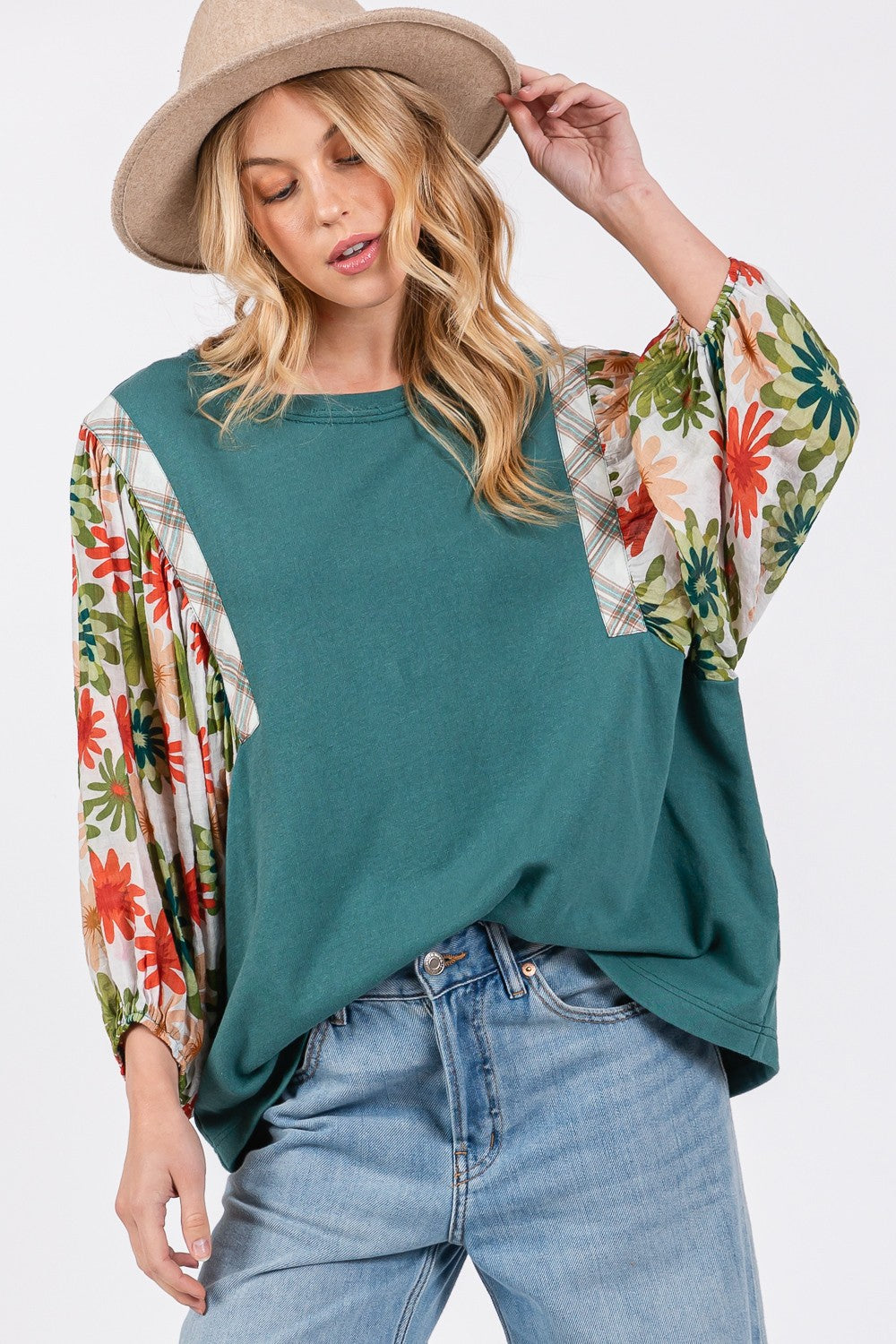 A person in a hat wears the SAGE + FIG Full Size Printed Balloon Sleeve Contrast Top with teal floral-patterned sleeves, blue jeans, and a back heart patch detail.