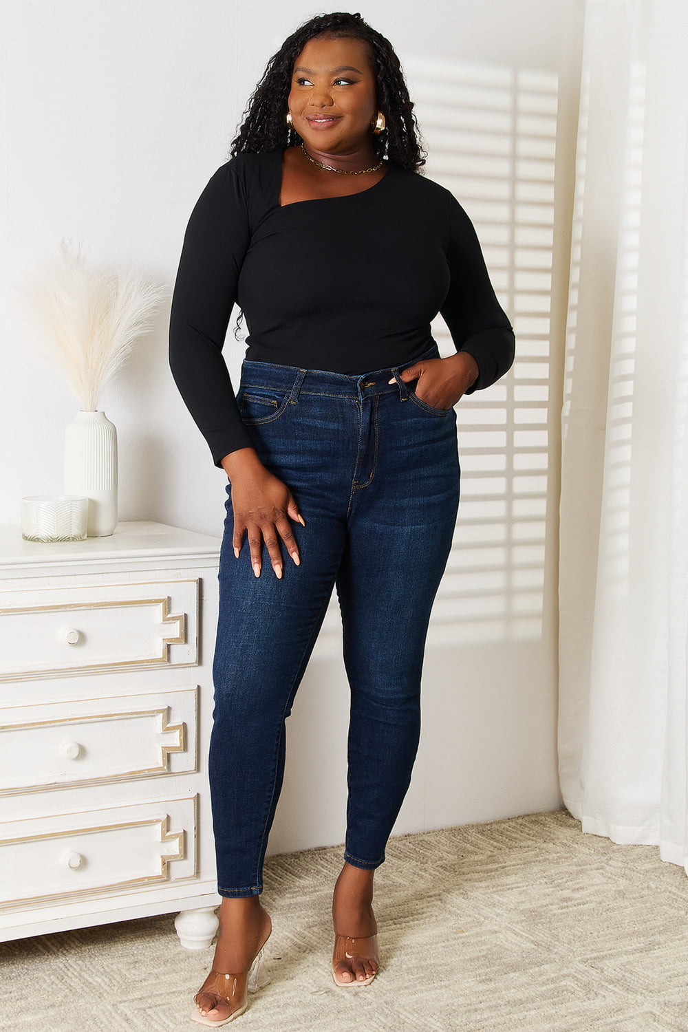 A woman with long hair is wearing a modern, stylish wardrobe featuring the Basic Bae Asymmetrical Neck Long Sleeve Top and jeans. She stands indoors in front of a white wall with shadow patterns.