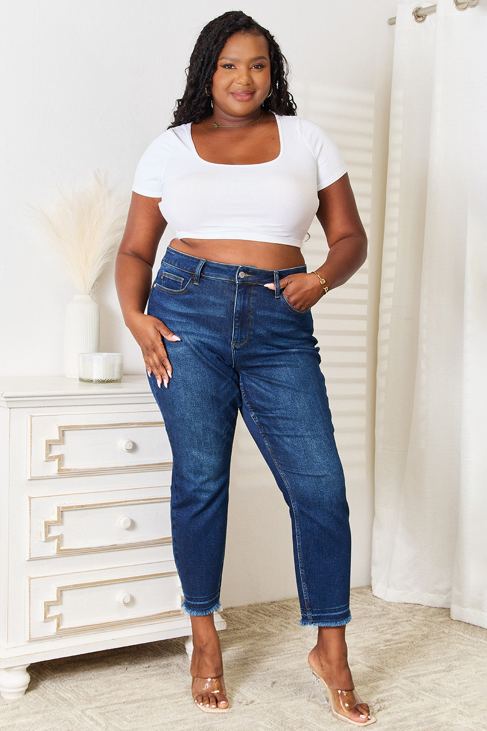 A person wearing Judy Blue Full Size High Waist Released Hem Slit Jeans in indigo wash and a white cropped top stands by a white dresser, showcasing a flattering fit.