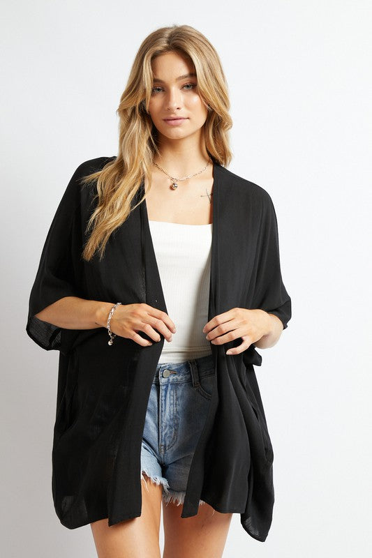 A woman poses against a simple backdrop, gracefully wearing a wardrobe essential: the Favorite Solid Kimono Cardigan in pink. Complementing her white tank top and denim shorts, this casual layer enhances her relaxed style with an elegant touch.