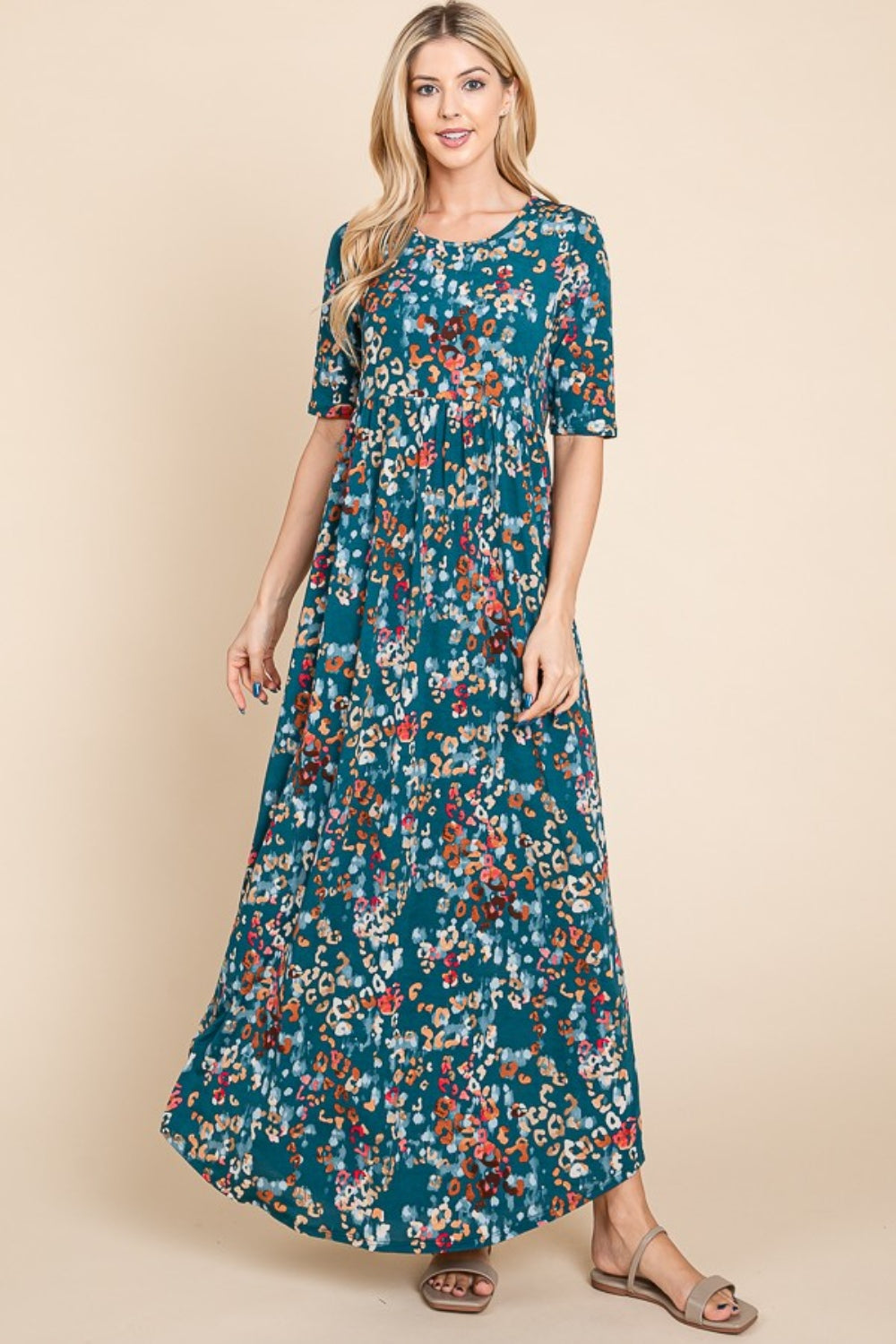 A person wearing a BOMBOM Printed Shirred Maxi Dress, featuring an eye-catching teal floral pattern and short sleeves, stands against a beige background.