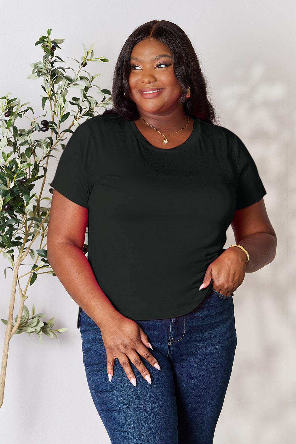 A woman with long hair, smiling with her right hand in her pocket and standing next to a plant, is wearing the Basic Bae Full Size Round Neck Short Sleeve T-Shirt in black along with beige pants made of rayon spandex.