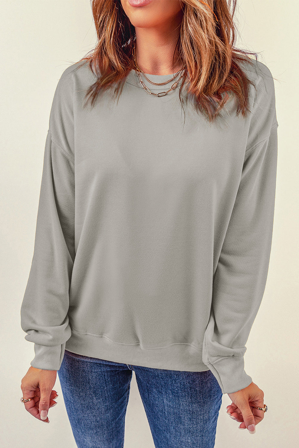 Person wearing a Gray Plain Crew Neck Pullover Sweatshirt and blue jeans with a hand in their pocket, adorned with layered necklaces.