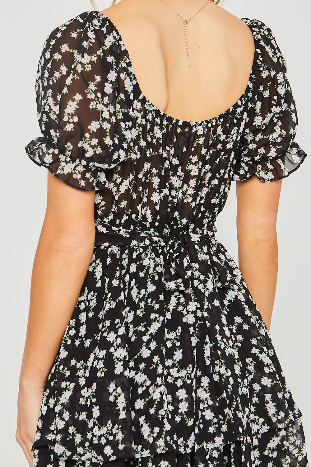 A woman is showcasing summer fashion in a short black floral romper from Love Tree, standing against a plain light background.