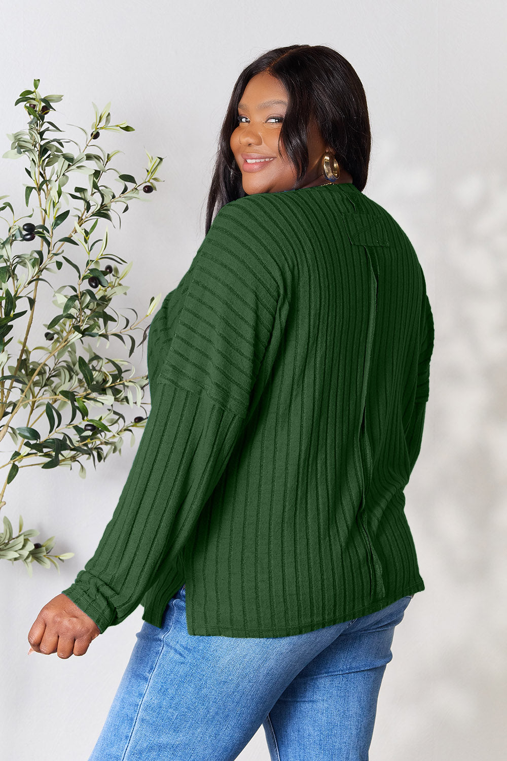 A person wearing the Basic Bae Full Size Ribbed Round Neck Slit Knit Top in blue and beige pants stands and smiles in front of a leafy plant.