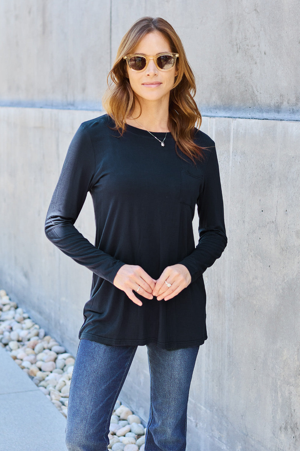 A woman with long hair wearing sunglasses, a Basic Bae Full Size Round Neck Long Sleeve Top in turquoise, and jeans is standing in front of a concrete wall with one hand on her hip.