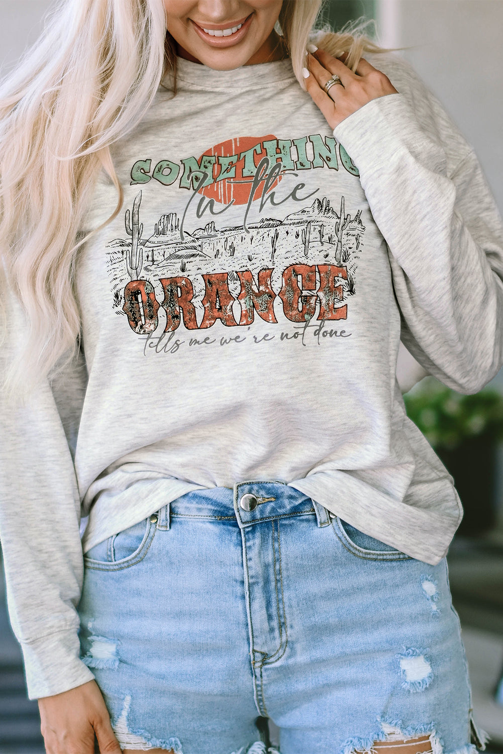 A person with long blonde hair is seen from behind, wearing denim shorts and a Gray SOMETHING ORANGE Graphic Relaxed Sweatshirt featuring a desert sunrise print.