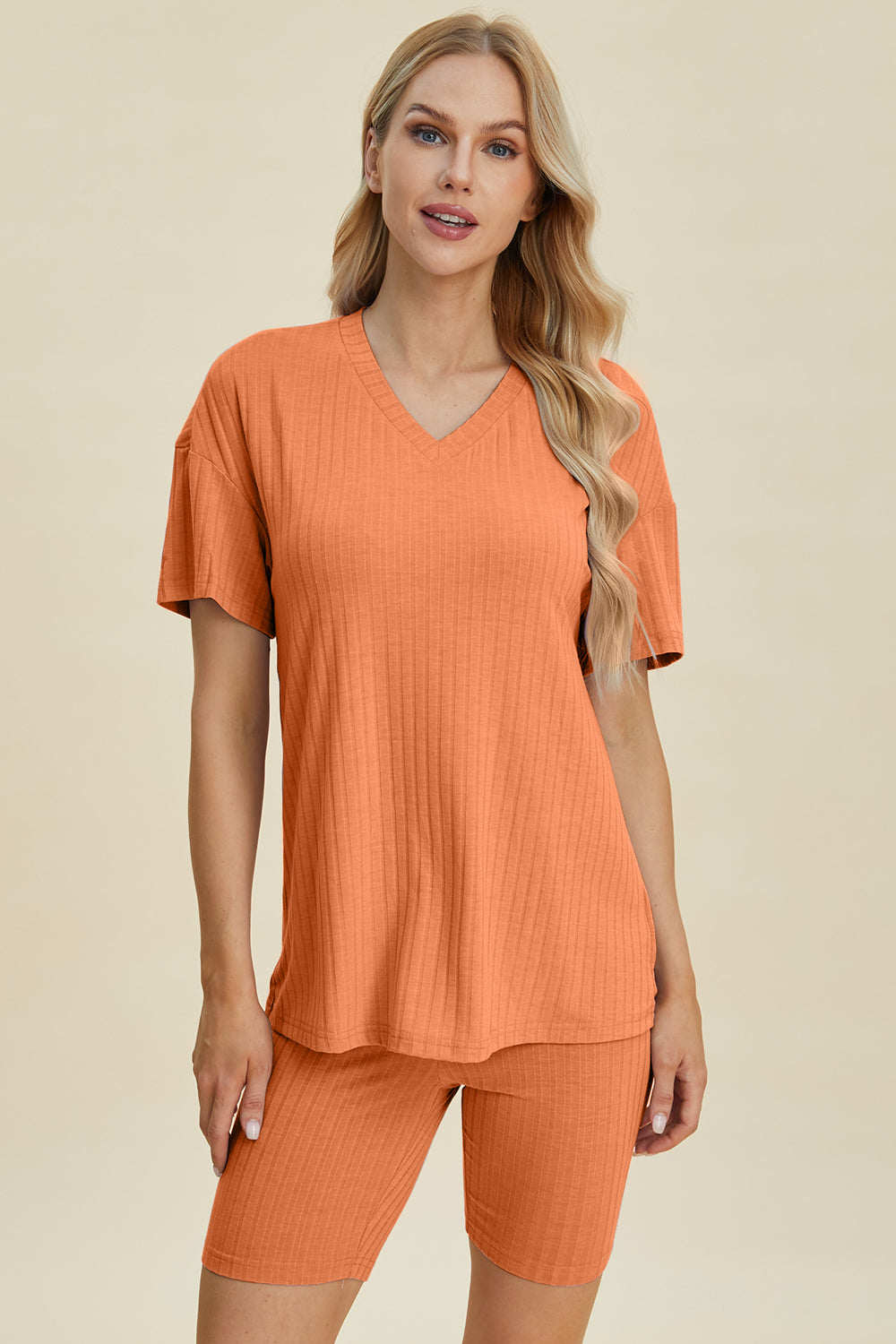 A woman with long blonde hair wearing the Basic Bae Full Size Ribbed V-Neck Short Sleeve Top and Shorts Set in rust stands against a plain background. This stylish two-piece set is made from a comfortable, stretchy fabric that is also machine washable.