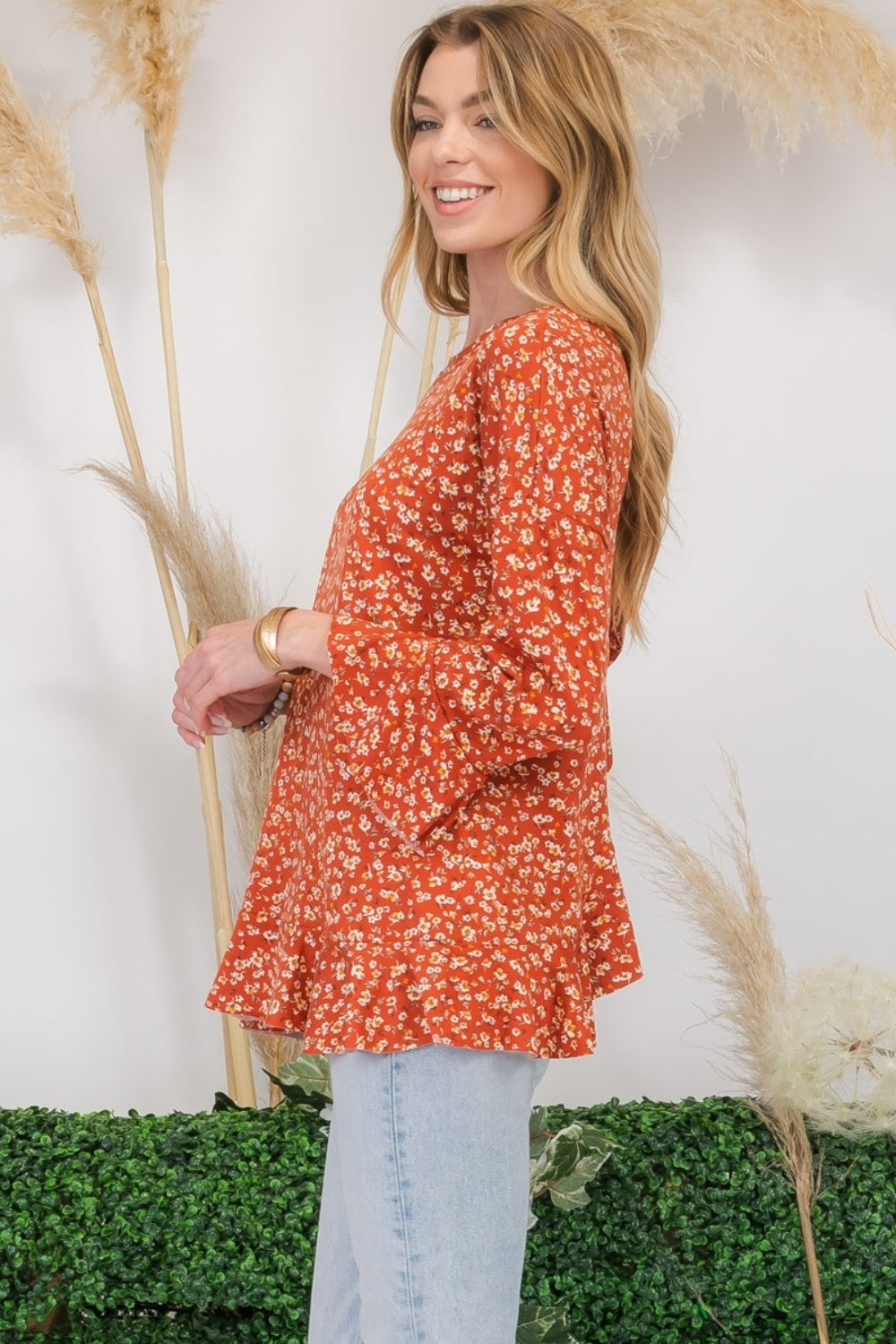 A woman wearing the Celeste Full Size Floral Ruffle Detail Top and jeans stands smiling in front of pampas grass and greenery, exuding feminine flair.