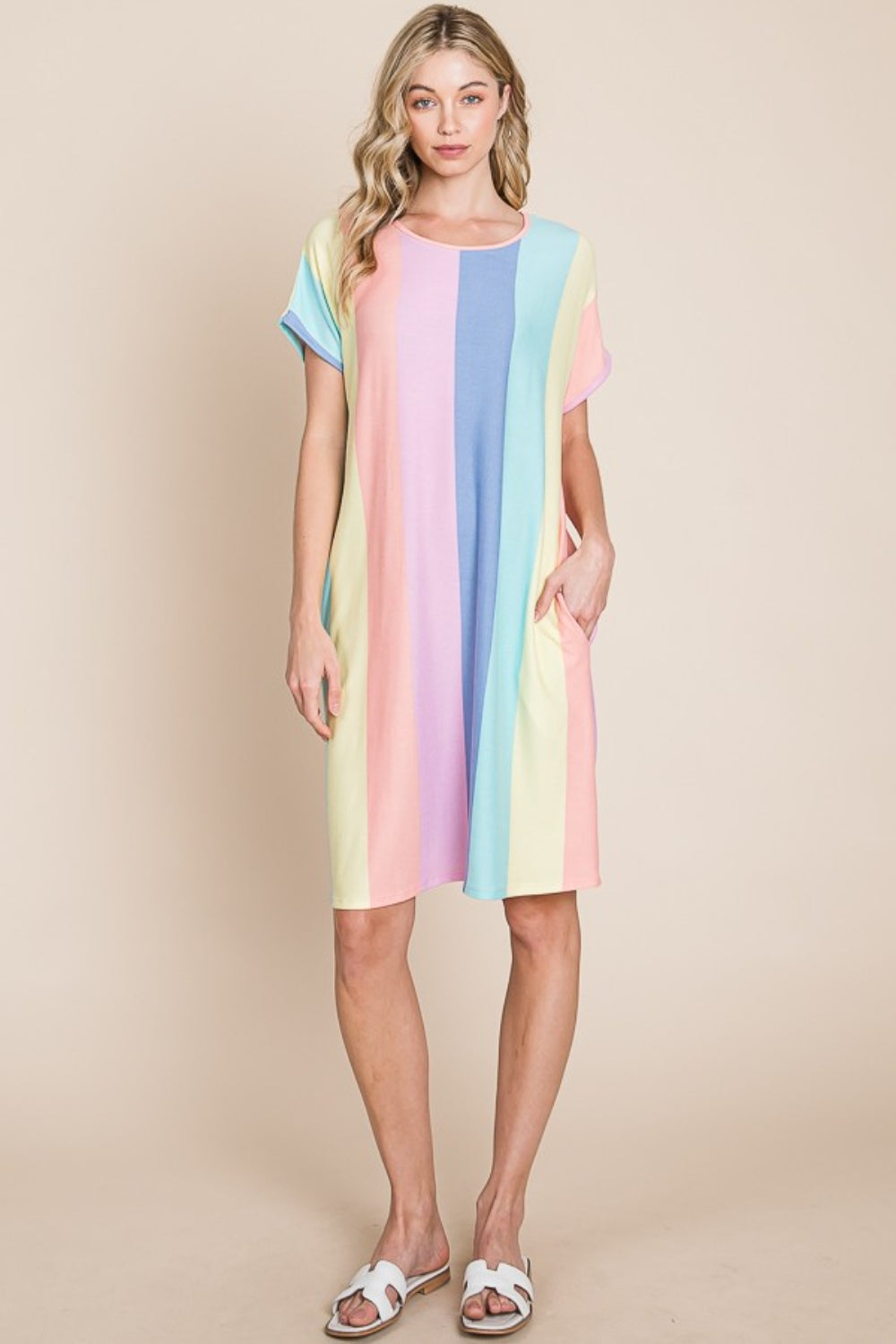 A person stands against a plain background, wearing the BOMBOM Striped Short Sleeve Dress with Pockets, crafted from comfortable fabric and featuring pastel stripes, with hands casually tucked into the convenient pockets.