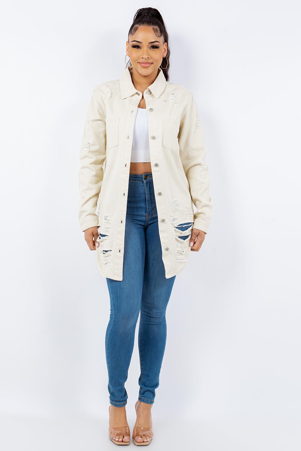 A woman wearing an American Bazi Distressed Button Up Long Sleeve Denim Jacket in beige, paired with a button-up white crop top and blue jeans, stands against a plain background.