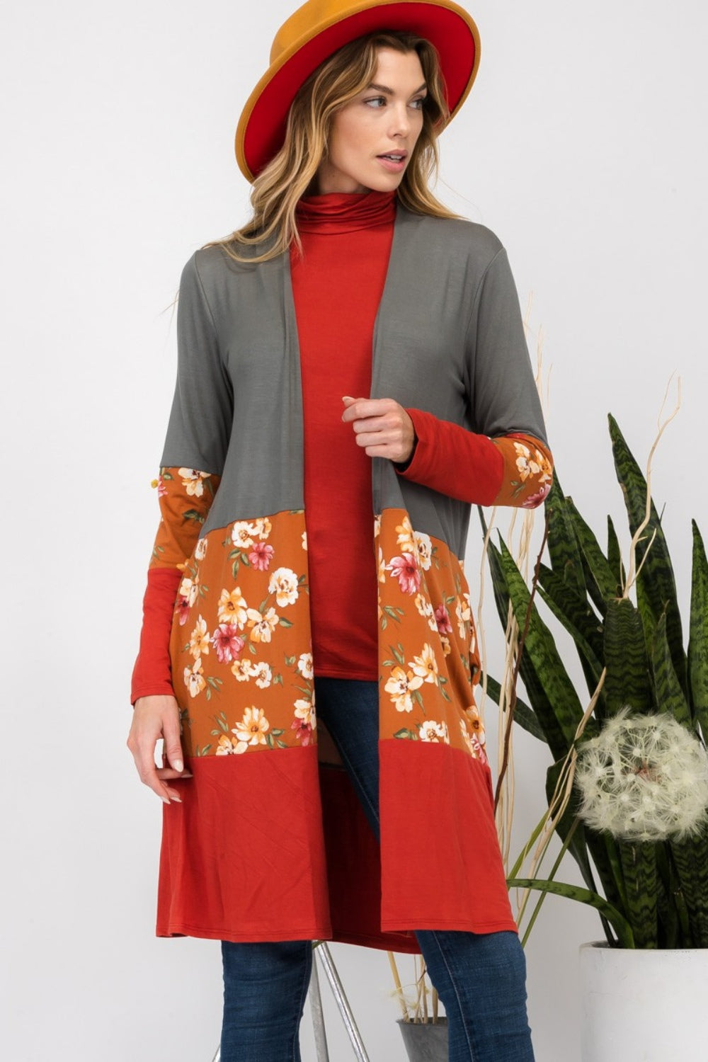 Wearing the Celeste Full Size Floral Color Block Open Front Cardigan, a person in a red hat stands indoors near potted plants, smiling.