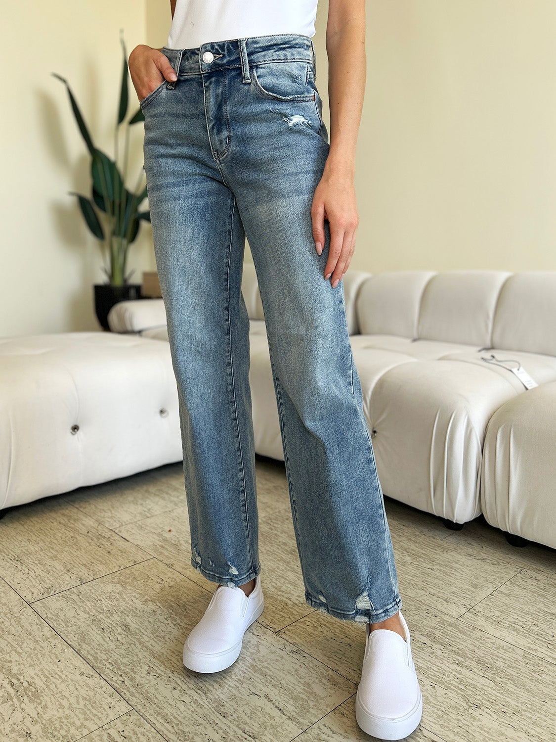 A person wearing Judy Blue Full Size Mid Rise Distressed Straight Jeans in light blue with a white top and white slip-on shoes stands in a room with a white couch and a plant.