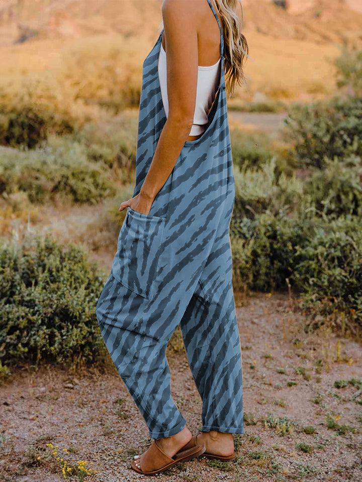 A person wearing a casual white V-neck tank top and a Full Size Printed V-Neck Sleeveless Jumpsuit with a brown animal print zebra-stripe pattern stands outdoors with one hand on their hat and the other in their pocket, set against a rocky landscape backdrop.