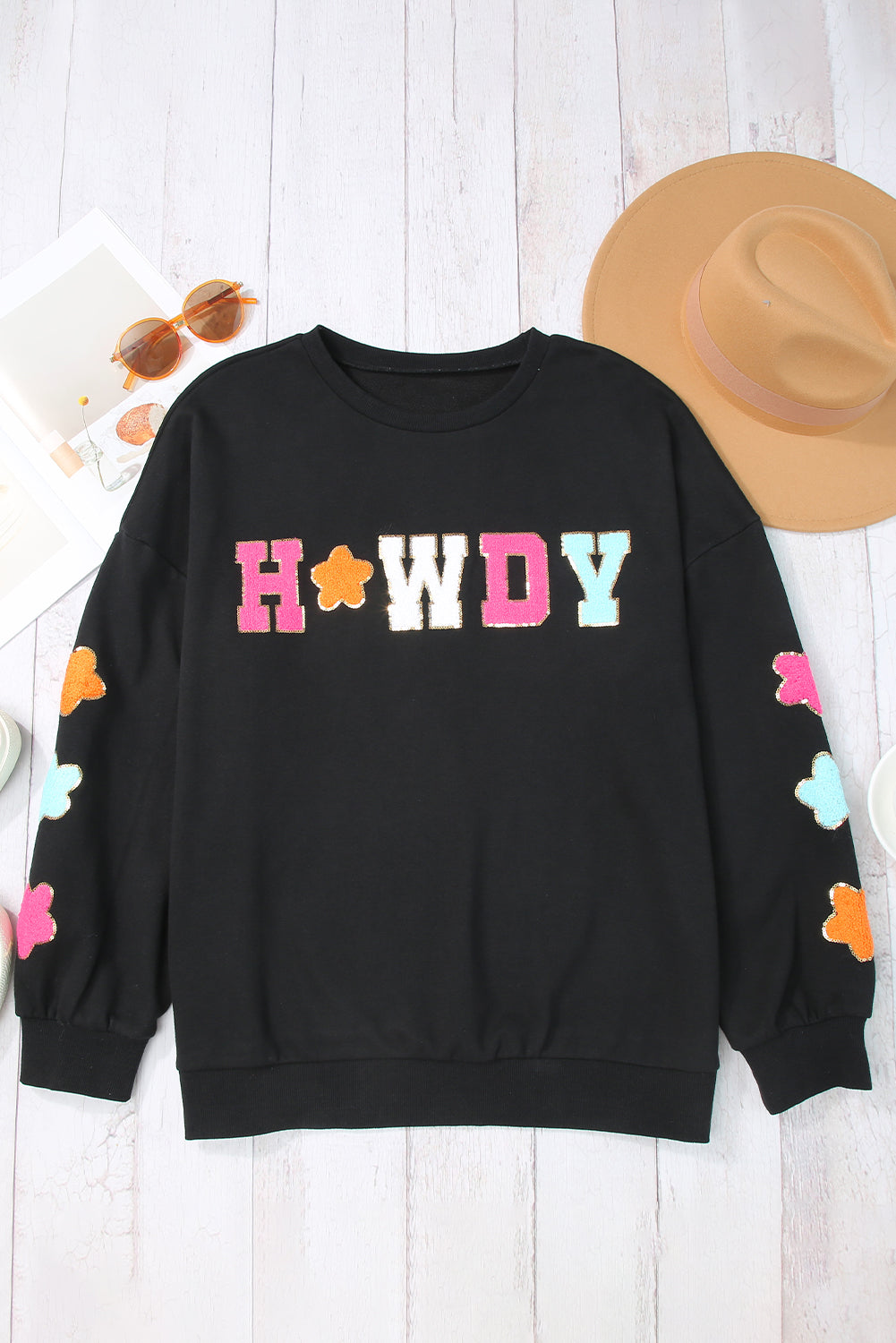 A woman with short blonde hair is wearing the Black Howdy Glitter Chenille Patch Graphic Casual Sweatshirt, featuring colorful star patterns and "WD" letters. She pairs it with light denim shorts and smiles while looking to the side.