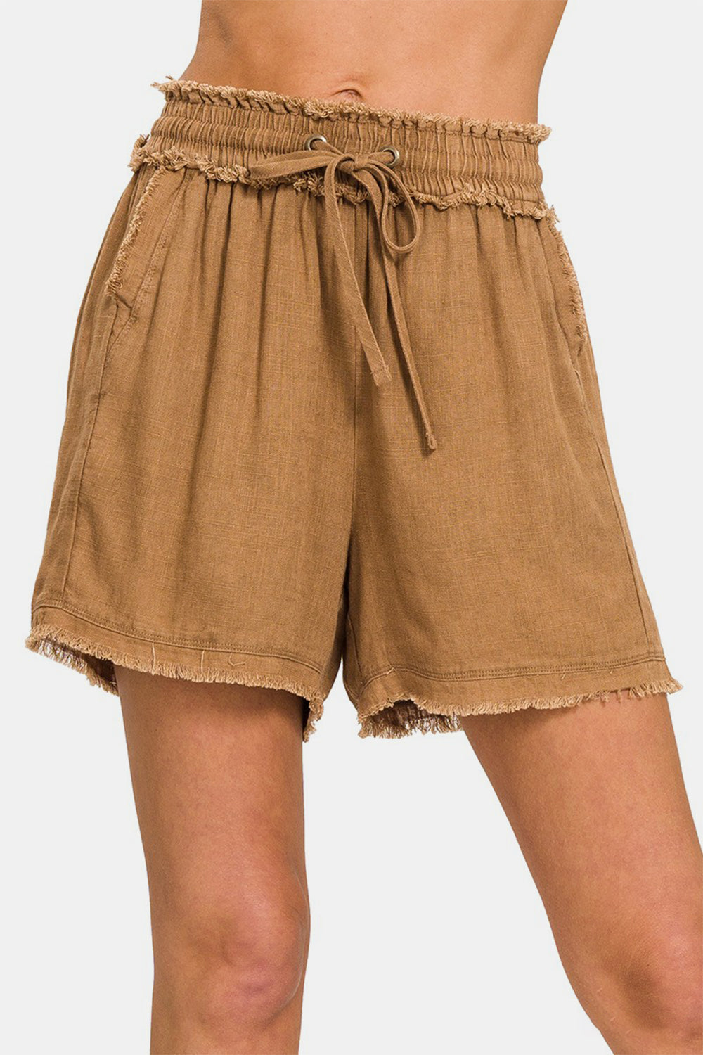 A person is wearing the Zenana Washed Linen Frayed Hem Drawstring Shorts, which come in a brown shade. These high-waisted shorts have a drawstring waist, frayed hem, and practical side pockets.