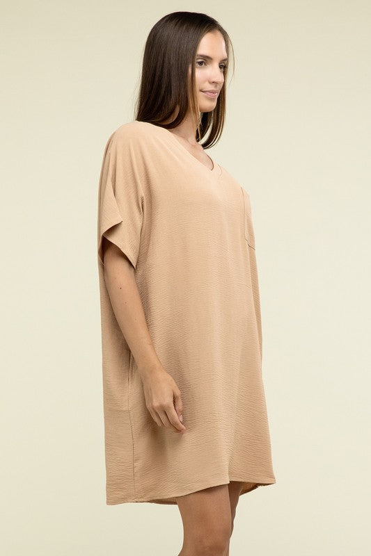 A woman wearing a loose light brown Woven Airflow V Neck T-Shirt Dress with Pockets stands against a plain background. Made from breathable fabric, this summer dress includes pockets where her hands rest as she smiles slightly.