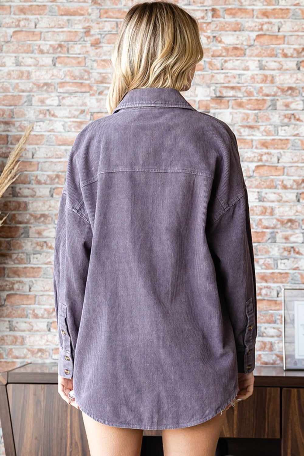 A blonde woman wearing the First Love Washed Corduroy Button Down Long Sleeve Shirt in purple over a white crop top and denim shorts stands in front of a brick wall.