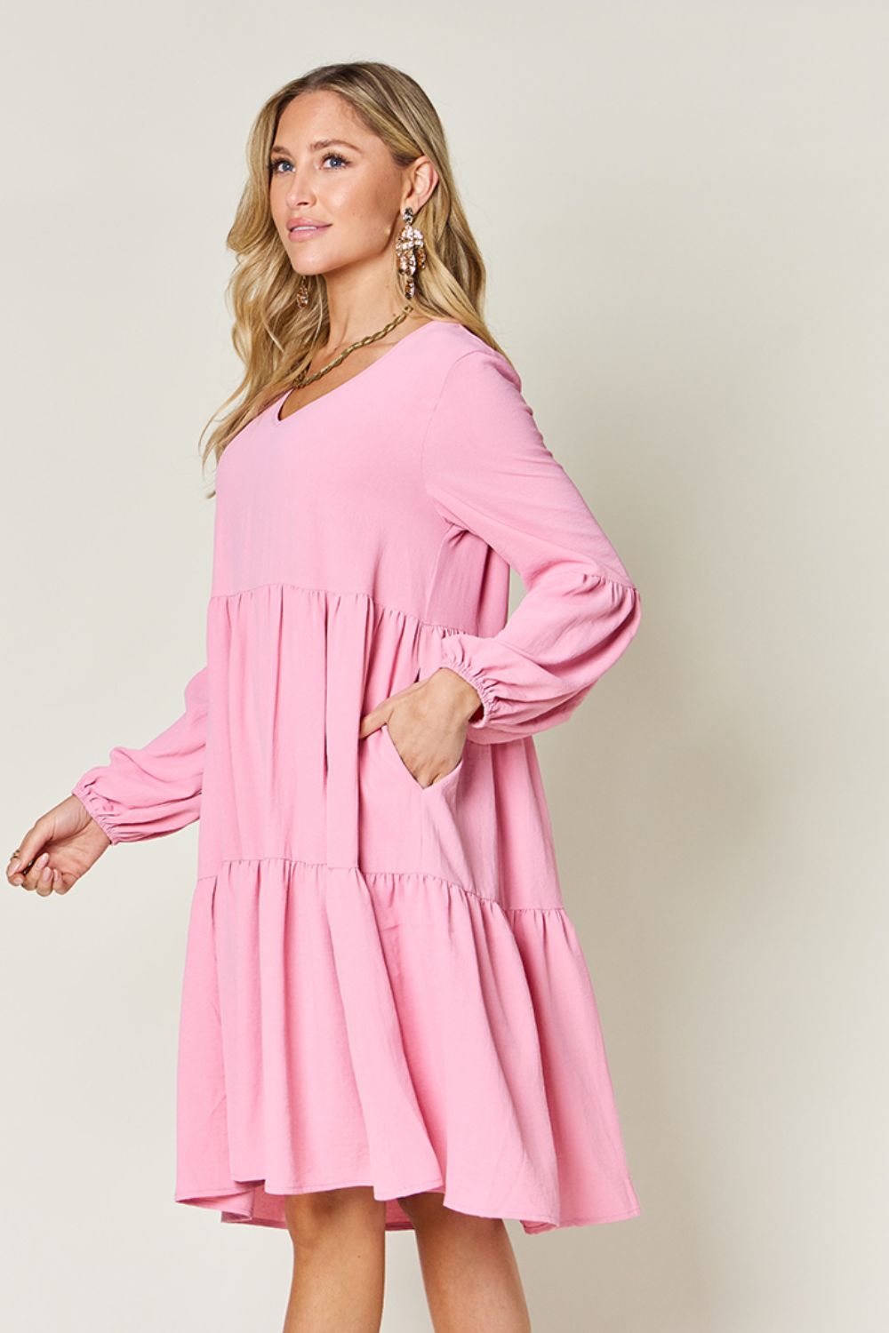 A woman stands wearing the Double Take Full Size V-Neck Balloon Sleeve Tiered Dress with Pockets in pink. Crafted from high stretch yarn, this dress combines comfort and style effortlessly. She has long, wavy hair and accessorizes with hoop earrings and a necklace. This imported garment is easy to care for – simply machine wash cold to keep it looking fresh.