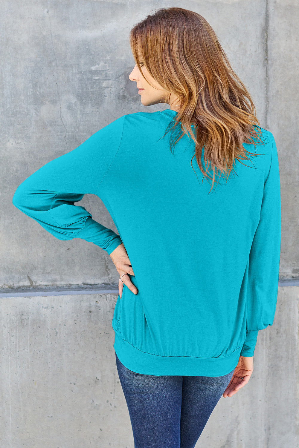 A woman with brown hair is standing against a concrete wall, wearing the Basic Bae Full Size V-Neck Lantern Sleeve Top in teal and blue basic-style jeans, smiling.