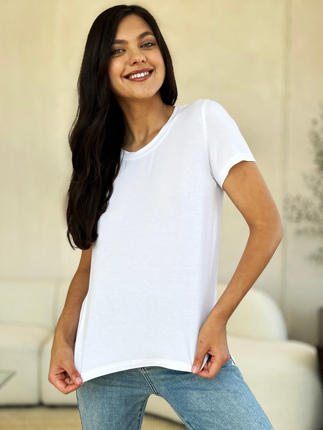 A woman stands next to an indoor plant, smiling, with one hand in her khaki pants pocket. In her pink Basic Bae Full Size Round Neck Short Sleeve T-Shirt and gold jewelry, she radiates casual elegance.