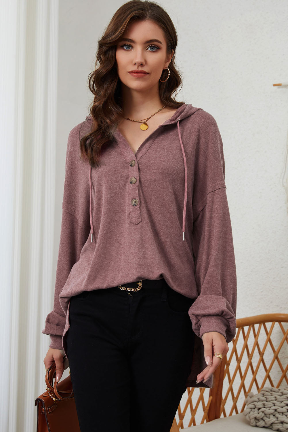 A woman stands facing away, wearing a stylish Buttoned High and Low Hem Hoodie in mauve made of high-quality fabric and black pants. She is in a room with light curtains and a wicker chair.